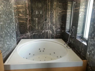 Corner bath tub beside a window with dark stone wall finishes, photograph published in Raj Plumbing's gallery